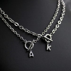 May include: A silver-toned double chain necklace featuring two toggle clasps as focal points. One toggle clasp is adorned with a small letter A charm, while the other toggle clasp features a small letter K charm, both suspended from the metal links.
