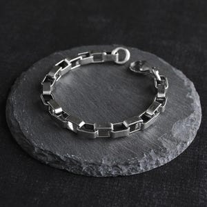 May include: A silver-colored bracelet with a rectangular link design rests on a dark gray, round stone. The bracelet is made of metal and has a clasp closure. The background is a dark, textured surface.