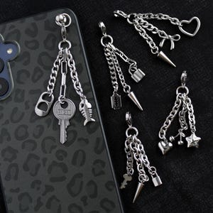 Op de afbeelding: Zilverkleurige telefoonbedels met diverse bungelende bedels, waaronder een sleutel, vis en flesopener. Andere bedels zijn harten, sterren en spikes. De telefoonhoes heeft een zwart-grijze luipaardprint.