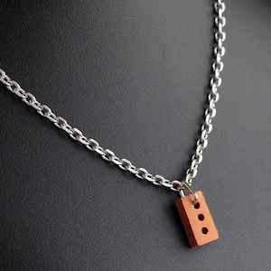 Könnte beinhalten: Halskette mit silberner Kette und einem kleinen, rechteckigen Anhänger in Form eines Ziegels. Der Ziegelanhänger ist rotbraun und hat drei runde Löcher. Die Halskette wird vor einem dunklen Hintergrund präsentiert.