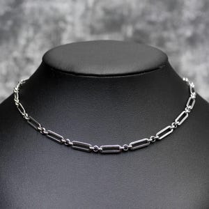 May include: A silver-colored chain necklace with elongated rectangular links, displayed on a black mannequin. The necklace has a delicate, minimalist design, suitable for everyday wear. The background is a mottled gray.