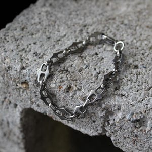 Op de afbeelding: Een zilverkleurige armband met een uniek kettingontwerp, rustend op een getextureerd grijs oppervlak. De schakels van de armband hebben een ingewikkelde, geometrische vorm en de sluiting is een simpele ring.