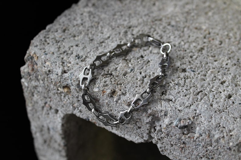 Op de afbeelding: Een zilverkleurige armband met een uniek kettingontwerp, rustend op een getextureerd grijs oppervlak. De schakels van de armband hebben een ingewikkelde, geometrische vorm en de sluiting is een simpele ring.
