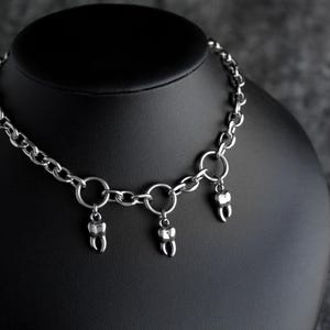 May include: A silver necklace featuring a chain and three tooth-shaped pendants. The necklace is displayed on a black mannequin against a dark grey background. The teeth are detailed with visible roots and enamel.