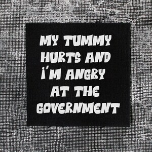 May include: A square black fabric patch featuring white embroidered text that reads: MY TUMMY HURTS AND I'M ANGRY AT THE GOVERNMENT. The lettering is in a bold, sans-serif font, and the patch has visible fabric edges against a textured background.