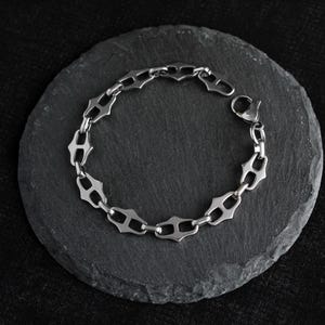 Op de afbeelding: Een zilverkleurige armband met een uniek schakelontwerp, rustend op een donkergrijze, cirkelvormige steen. De armband heeft een reeks gestileerde, open schakels, die een ketting vormen. De achtergrond is een donker, getextureerd oppervlak.