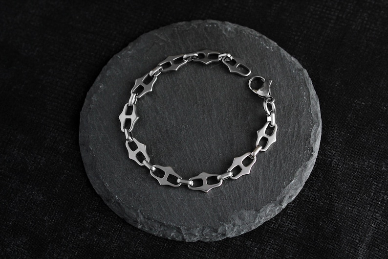 Op de afbeelding: Een zilverkleurige armband met een uniek schakelontwerp, rustend op een donkergrijze, cirkelvormige steen. De armband heeft een reeks gestileerde, open schakels, die een ketting vormen. De achtergrond is een donker, getextureerd oppervlak.