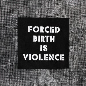 Könnte beinhalten: Ein quadratischer schwarzer Stoffaufnäher mit dem weißen, schablonenartigen Text FORCED BIRTH IS VIOLENCE in der Mitte. Die Schrift verwendet eine fette, blockartige Schriftart mit abgenutztem Aussehen für ein kontrastreiches Design.