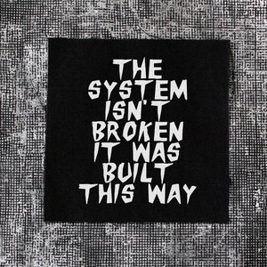 May include: A black square patch with white text that reads: "THE SYSTEM ISN'T BROKEN IT WAS BUILT THIS WAY." The patch is set against a textured, gray and white background.
