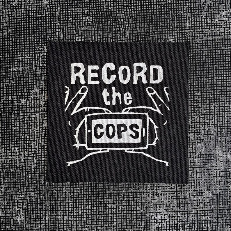 May include: Black and white patch with the text "RECORD the COPS" and a drawing of hands holding a phone.