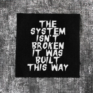 Puede incluir: Un parche de tela cuadrado de color negro con el texto en blanco THE SYSTEM ISN'T BROKEN IT WAS BUILT THIS WAY en una tipografía negrita, irregular y de estilo dibujado a mano. El texto está centrado y ocupa la mayor parte del parche.