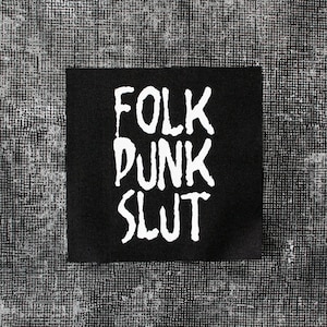 May include: A black square patch featuring the words "FOLK PUNK SLUT" in white, handwritten-style lettering. The patch is set against a textured, grey and white background.