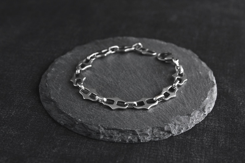 Op de afbeelding: Een zilverkleurige armband met een uniek schakelontwerp, rustend op een donkergrijze, cirkelvormige steen. De schakels van de armband hebben een gestileerde, geometrische vorm. De achtergrond is een donker, getextureerd oppervlak.