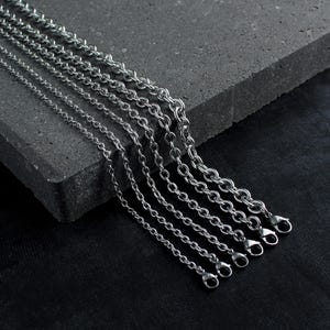 May include: A collection of six silver-toned metal chains of varying thicknesses, each featuring a polished finish and a secure lobster claw clasp at the end. The chains are arranged in a parallel row, showcasing different link sizes from thin to thick.