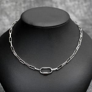 May include: A silver-colored chain necklace with a rectangular link design. The necklace features a large oval clasp in the center. The jewelry is displayed on a black mannequin head.