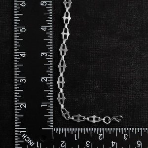 Op de afbeelding: Een zilverkleurige kettingarmband met een uniek schakelontwerp, ongeveer 15 cm lang. De armband wordt getoond tegen een zwarte achtergrond met een liniaal voor de schaal. De schakels hebben een opvallende vorm.