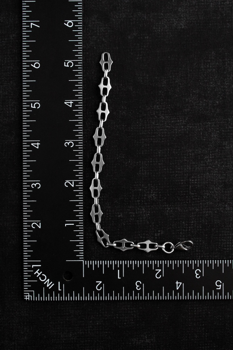 Op de afbeelding: Een zilverkleurige kettingarmband met een uniek schakelontwerp, ongeveer 15 cm lang. De armband wordt getoond tegen een zwarte achtergrond met een liniaal voor de schaal. De schakels hebben een opvallende vorm.