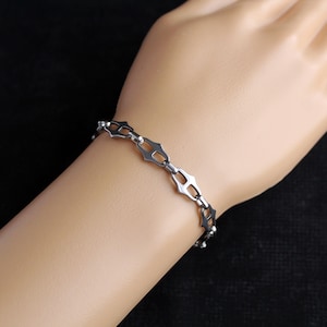Op de afbeelding: Een zilverkleurige armband met een geometrisch ontwerp. De armband heeft een reeks onderling verbonden, open gevormde schakels. De armband wordt op de arm van een persoon tegen een donkere achtergrond getoond. De schakels zijn ongeveer 1,3 cm lang.
