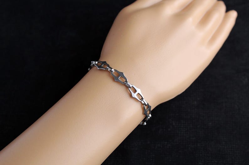 Op de afbeelding: Een zilverkleurige armband met een geometrisch ontwerp. De armband heeft een reeks onderling verbonden, open gevormde schakels. De armband wordt op de arm van een persoon tegen een donkere achtergrond getoond. De schakels zijn ongeveer 1,3 cm lang.