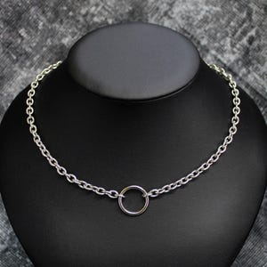 Op de afbeelding: Een zilverkleurige ketting met een cirkelvormige hanger. De ketting wordt gepresenteerd op een zwarte mannequinhoofdstuk, tegen een grijze achtergrond. De schakels van de ketting zijn ovaal van vorm.