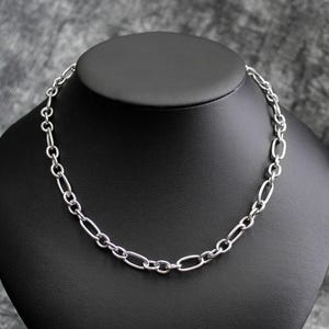 May include: A silver-colored chain necklace with oval and round links is displayed on a black mannequin. The necklace is a choker style, with a simple and elegant design. The background is a textured gray.