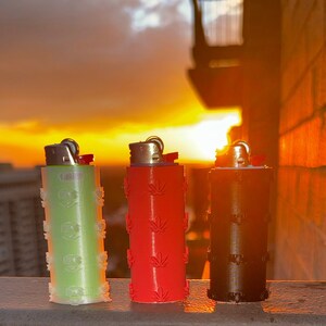 Puede incluir: Tres encendedores con diseños de diferentes colores. Los encendedores son verde, rojo y negro. Los encendedores son todos del mismo tamaño y forma. Están sentados en una barandilla con una puesta de sol en el fondo.