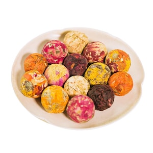 May include: A collection of colorful, spherical tea balls on a white plate. The tea balls are composed of dried flowers in shades of orange, yellow, pink, and brown. The plate has a slightly raised edge and a neutral color.