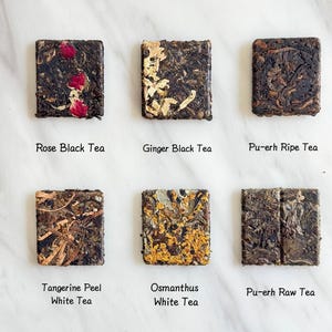 May include: Overhead shot of six square tea cakes. The cakes are labeled: Rose Black Tea, Ginger Black Tea, Pu-erh Ripe Tea, Tangerine Peel White Tea, Osmanthus White Tea, and Pu-erh Raw Tea. The tea cakes are various shades of brown and tan.