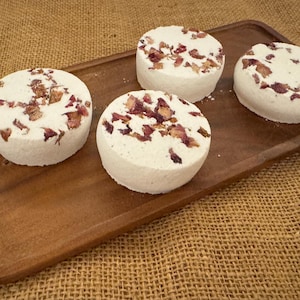 May include: Four round, white bath bombs with dried rose petals on a wooden tray. The bath bombs are arranged in a row, and the tray is a dark brown color. The background is a burlap texture.