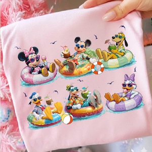 Puede incluir: Camiseta rosa claro con un gráfico colorido de personajes de Disney relajándose en flotadores inflables en el agua. Minnie, Mickey, Pluto, Donald, Goofy y Daisy disfrutan de bebidas tropicales y helados.
