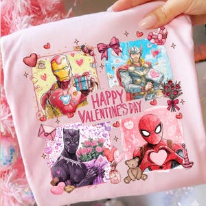 May include: A light pink t-shirt featuring four panels with Marvel characters Iron Man, Thor, Black Panther, and Spider-Man, each holding hearts or gifts. The words "HAPPY VALENTINE'S DAY" are printed in the center. Hearts and stars decorate the design.