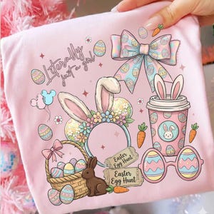May include: Pink graphic tee with Easter-themed illustrations. Features a bunny ear headband, coffee cup with bunny ears, Easter eggs, a bow, and the text "Literally just a girl." Includes a basket with eggs, a brown bunny, and carrot accents.