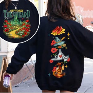 May include: Black sweatshirt with a colorful graphic from the Wizard of Oz. The front features text "OFF TO SEE THE WIZARD" above a yellow brick road leading to a green castle. The back has illustrations of ruby slippers, a witch's hat, and other themed elements.