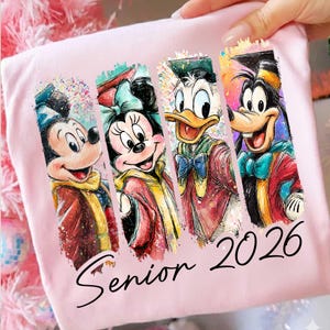 May include: A light pink t-shirt featuring four cartoon characters: Mickey Mouse, Minnie Mouse, Donald Duck, and Goofy, each in graduation attire. The words "Senior 2026" are printed in black script below the characters.