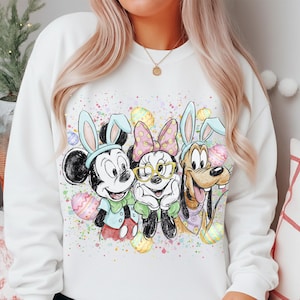 Puede incluir: Sudadera blanca con Mickey Mouse, Minnie Mouse y Pluto disfrazados de conejos de Pascua. El diseño incluye coloridos huevos de Pascua y un efecto de salpicaduras. Los personajes llevan orejas de conejo y sonríen. Un diseño festivo para la festividad de Pascua.