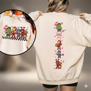 May include: Cream-colored sweatshirt featuring cartoon illustrations of Marvel superheroes. The front displays Hulk, Iron Man, Captain America, and Spider-Man with hearts and balloons. The back has a vertical design with the same characters and bows.