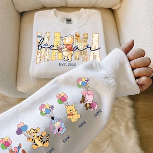 May include: White sweatshirt with "Mama Bear" text and Winnie the Pooh graphic. The sleeve features cartoon characters with balloons. The text "EST. 2026" is below the graphic. The sleeve has the word "name" below each character.