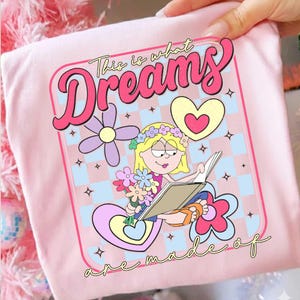 Op de afbeelding: Zachtroze T-shirt met een grafisch ontwerp. Het ontwerp bevat de tekst "This is what Dreams are made of", een stripmeisje dat leest, bloemen en harten in een retro-stijl.