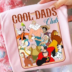Puede incluir: Camiseta rosa claro con un estampado de personajes de dibujos animados y el texto "COOL DADS Club" en marrón y azul. El diseño incluye personajes como Goofy, el Rey Tritón y Mr. Increíble.