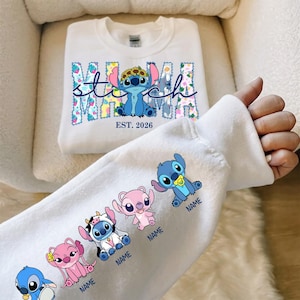 Könnte beinhalten: Weißes Sweatshirt mit einem blauen Stitch-Charakter, der einen Blumenkranz trägt, mit dem Wort "Mama" und "Stitch" in einem floralen Design. Der Ärmel zeigt mehrere Stitch-Charaktere. Der Text "EST. 2026" ist ebenfalls sichtbar.