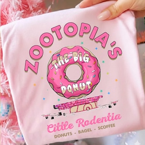 Könnte beinhalten: Hellrosa T-Shirt mit dem Schriftzug "Zootopia's" in Pink und einer pinken Donut-Grafik mit der Aufschrift "The Big Donut". Darunter eine pinke Fahrzeuggrafik sowie der Text "Little Rodentia" und "Donuts - Bagel - Scoffee".