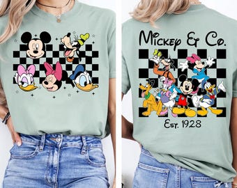 May include: Sage green t-shirt featuring Mickey Mouse, Donald Duck, Daisy Duck, Minnie Mouse, and Goofy on a black and white checkered background. The back of the shirt displays the text "Mickey & Co. Est. 1928" with additional characters.
