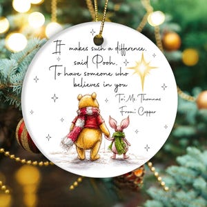 May include: A round, white Christmas ornament featuring Winnie the Pooh and Piglet holding hands. The ornament has a quote that reads "It makes such a difference, said Pooh. To have someone who believes in you." The ornament is personalized.