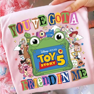 May include: A light pink t-shirt featuring a colorful graphic of various Toy Story characters surrounding a green frog-shaped frame. The center of the frame displays the text Disney Pixar Toy Story 5. The text You've Got A Friend In Me surrounds the design.