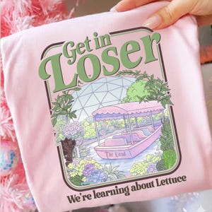 Peut inclure: T-shirt rose clair avec un motif graphique représentant une scène de promenade en bateau avec le texte "Get in Loser" et "We're learning about Lettuce". Le motif comprend un bateau, de l'eau et des éléments floraux.