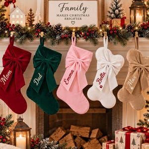 May include: Five velvet Christmas stockings hanging in a row. The first is red with 'Mom', the second green with 'Dad', the third pink with 'Emma', the fourth white with 'Bella' and a paw print, and the fifth tan with 'Max' and a paw print.
