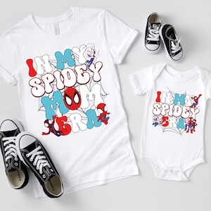 Könnte beinhalten: Weiße T-Shirts und ein Baby-Body mit dem Text "In My Spidey Era" in einem farbenfrohen Comic-Stil. Das Design umfasst Spider-Man-Charaktere und ein Spinnennetz. Schwarze Turnschuhe sind ebenfalls im Bild.
