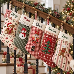 Op de afbeelding: Vijf gepersonaliseerde kerstsokken hangen aan een houten trapleuning. De sokken zijn voorzien van de geborduurde namen Riley, Mom, Dad, Amelia en Luna, met feestelijke ontwerpen zoals een teddybeer, sneeuwpop, notenkraker, kerstboom en peperkoekmannetje.