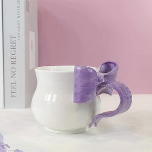 May include: White ceramic mug with a purple bow-shaped handle. The mug is sitting on a white surface with a stack of books in the background.