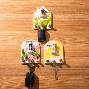 May include: Ceramic key holders with dog figurines. The key holders are white with colorful accents and feature the names "Peanut", "Liam & Emma", and "Charlie". Each holder has hooks for keys and decorative elements.